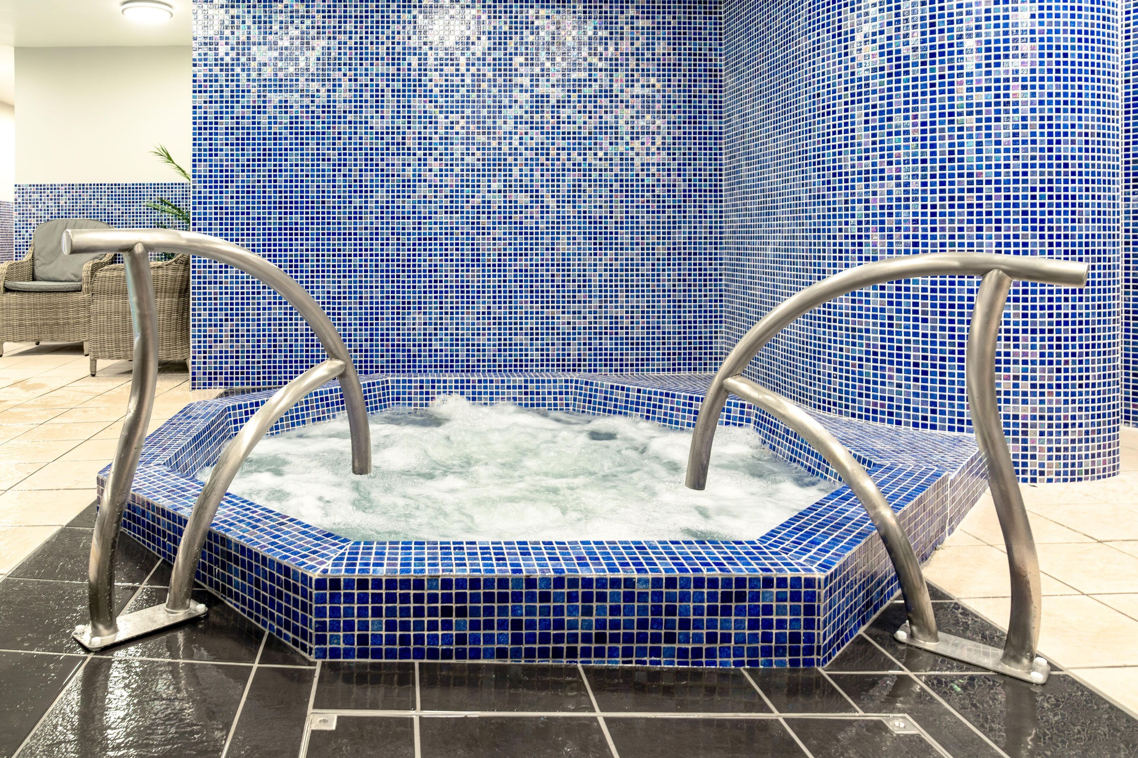 A hot tub with blue mosaic tiles and metal handrails, surrounded by a tiled floor and a wicker chair.