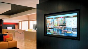 A modern hotel lobby with a large digital display on the wall, a reception desk, a comfortable seating area with a sofa and cushions, and a clean, tiled floor.