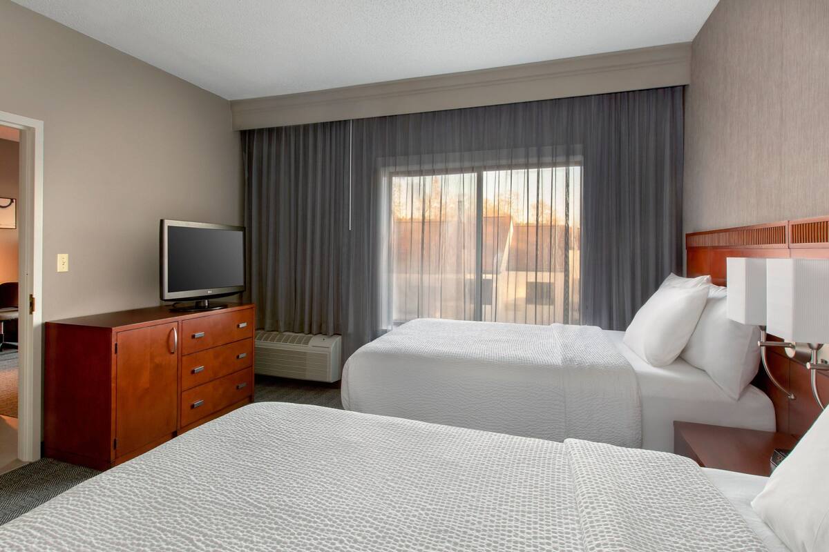 Una habitación de hotel con dos camas, televisión, cómoda y una ventana grande con cortinas.