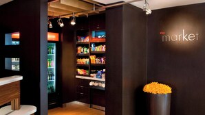 A modern interior with a dark wall, a wooden reception desk, a refrigerator stocked with beverages, shelves with snacks, and a large vase with orange flowers.