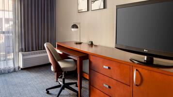 Quarto de hotel com escrivaninha de madeira, cadeira, televisão de tela plana, abajur e aquecedor. Há duas fotos emolduradas na parede.