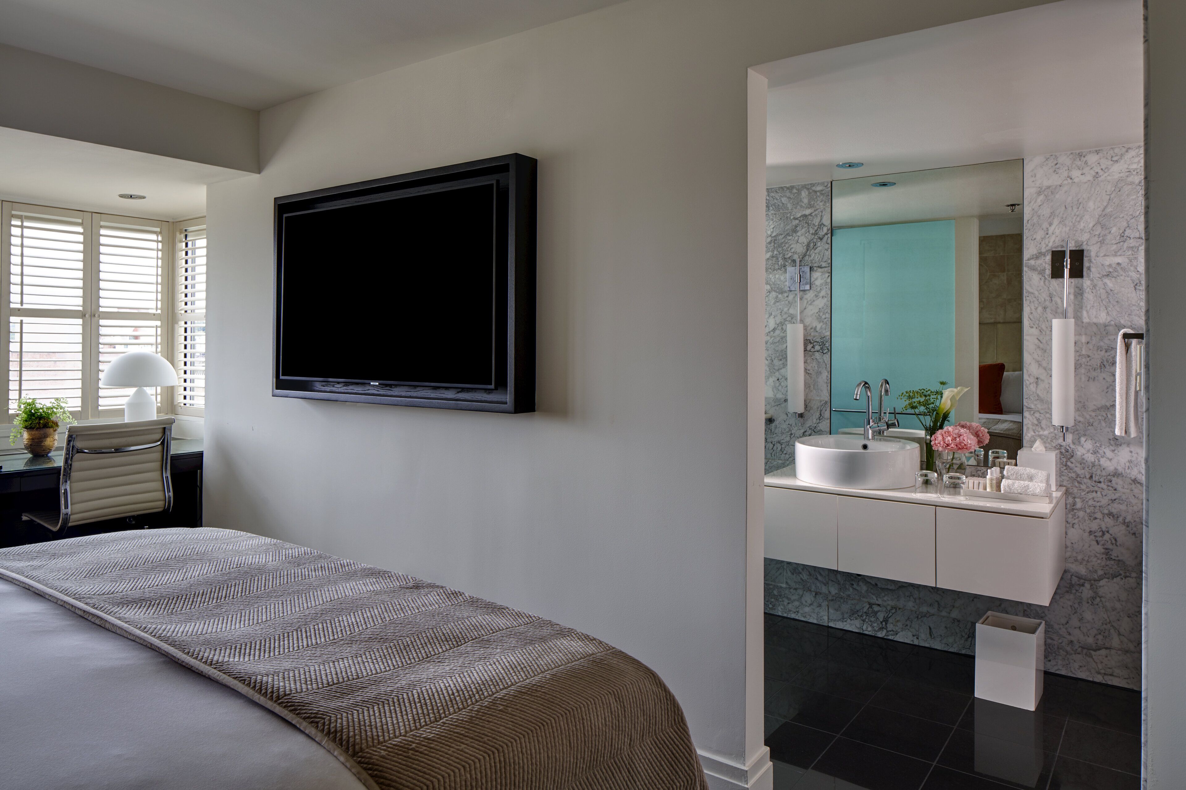 Een moderne slaapkamer met een groot bed, een wandgemonteerde televisie, en een badkamer met een marmeren wastafel en een grote spiegel.