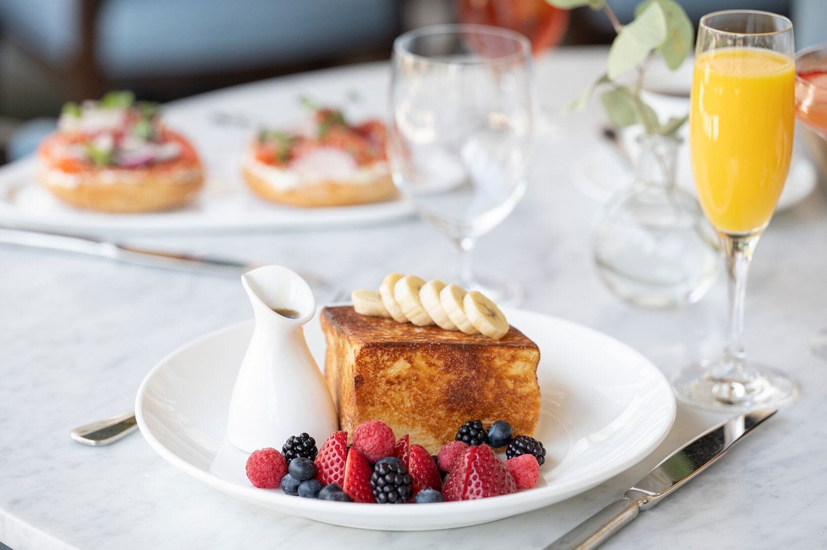 Ein Frühstückstisch mit Toast, belegt mit Bananenscheiben, Beeren und einer kleinen Kanne Sirup.
