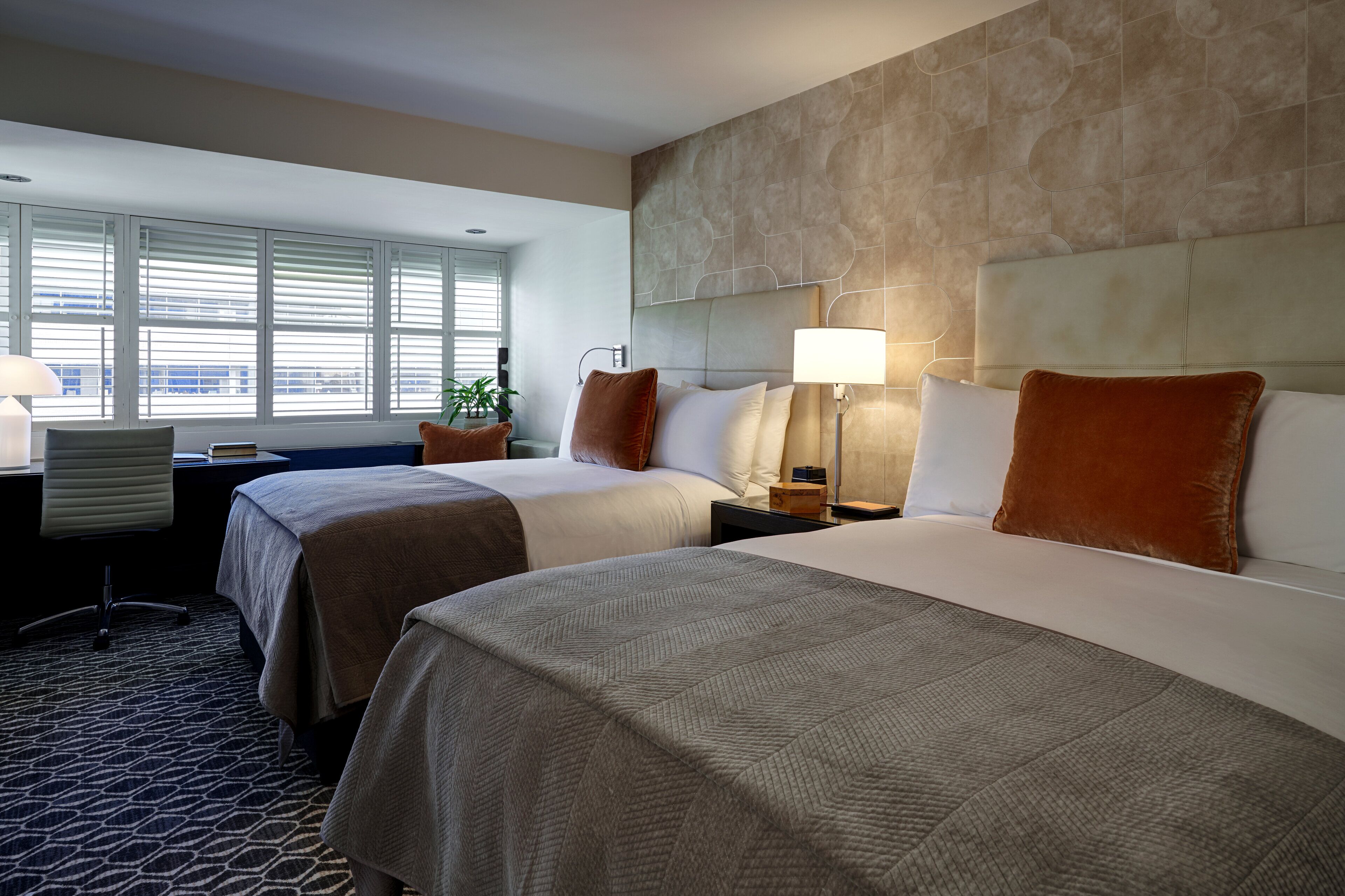 A hotel room with two beds, a desk, and a window with blinds.
