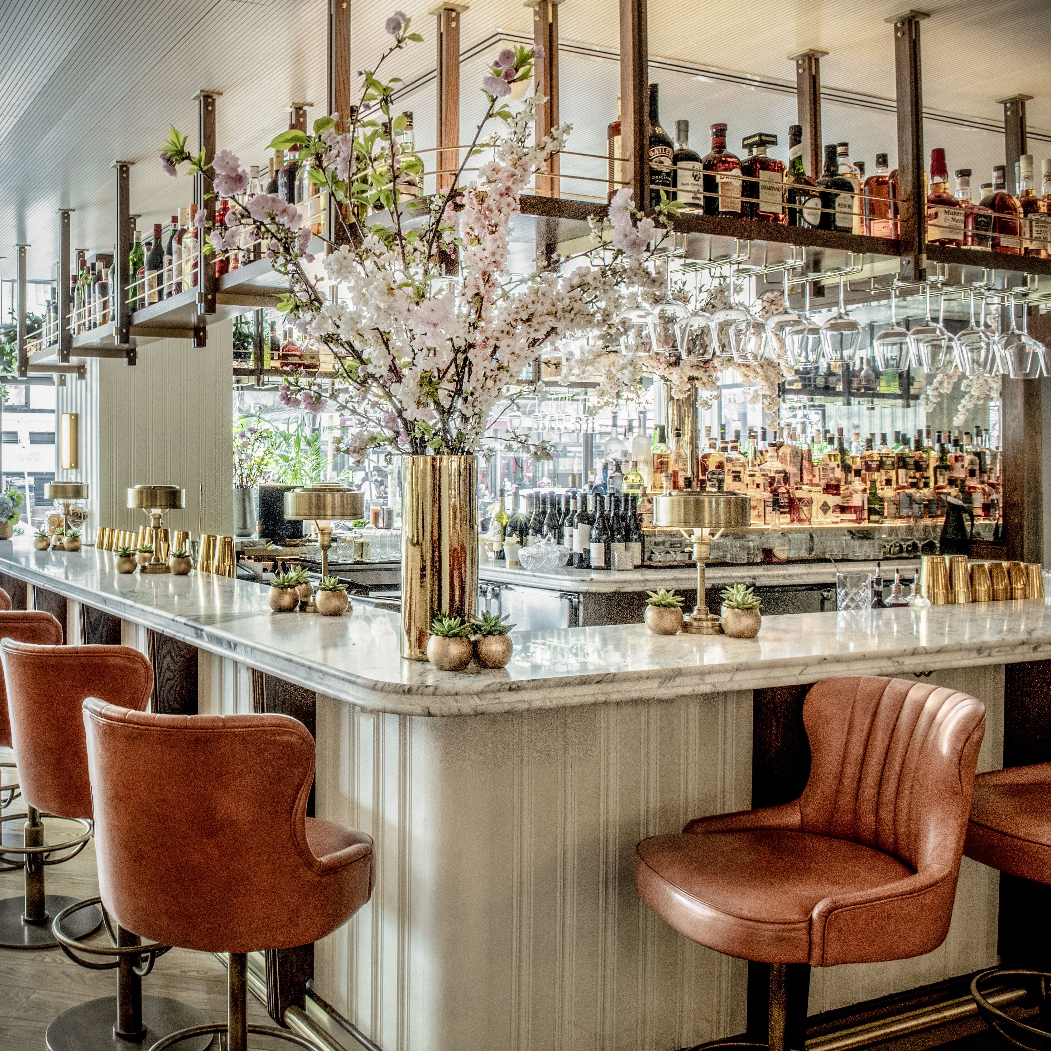 En bar med en marmorbordplade, mange flasker spiritus og en stor blomsteropsætning. Der er flere polstrede barstole og en høj, dekorativ vase.