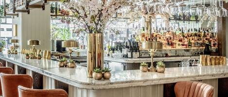 A bar area with a marble countertop, multiple liquor bottles, and a large floral arrangement. There are several cushioned bar stools and a tall, decorative vase.
