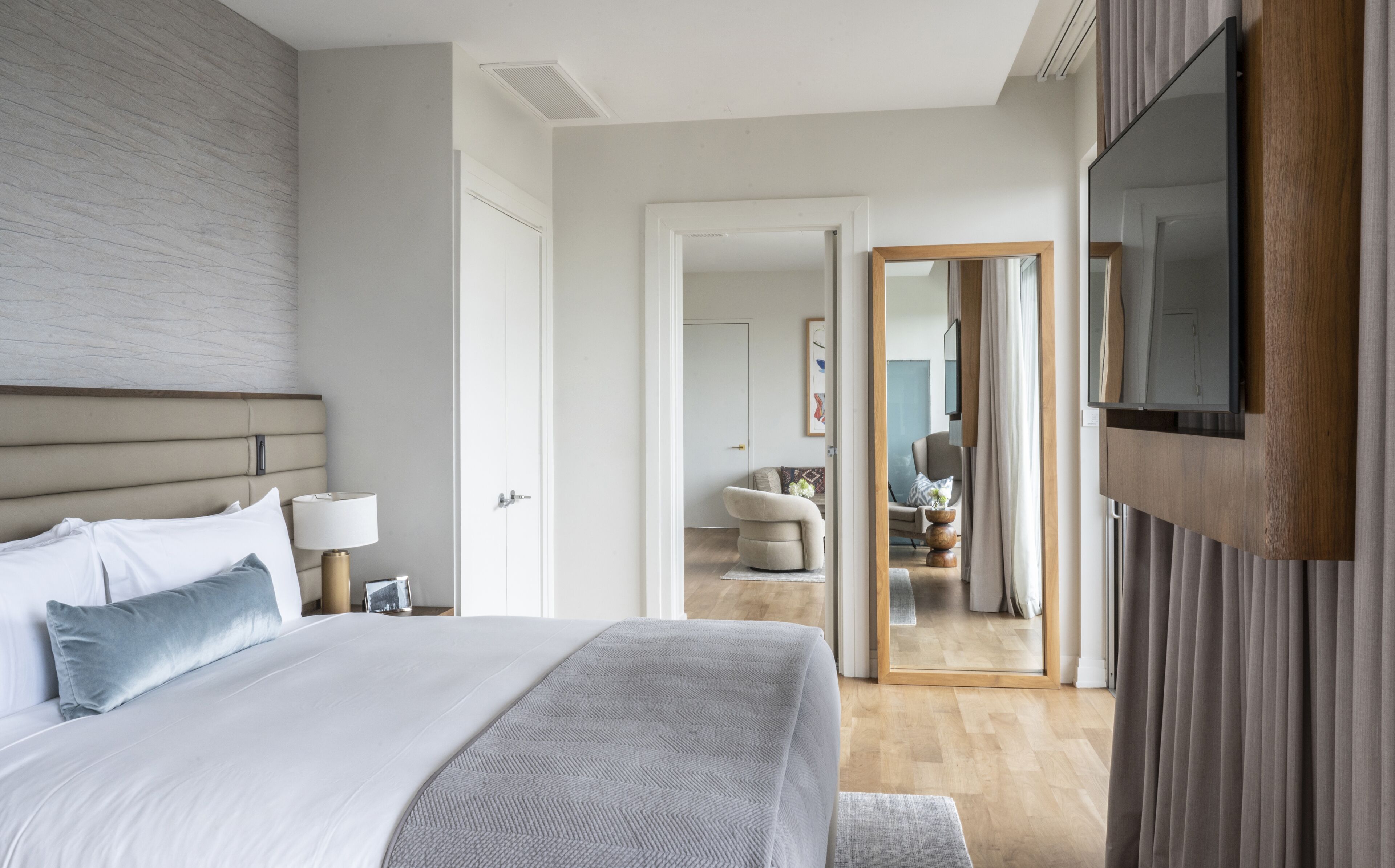 A modern bedroom with a large bed, a TV mounted on the wall, and a view into the adjacent living area.