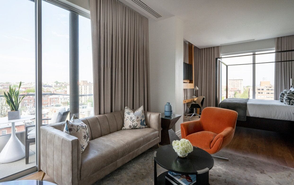 A modern hotel room with a large window, a sofa, an orange armchair, a bed, and a small table with a vase.