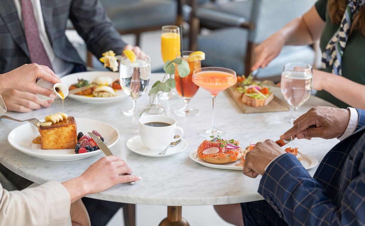 Een groep mensen dineert aan een ronde tafel met verschillende gerechten, drankjes en een kleine plant.