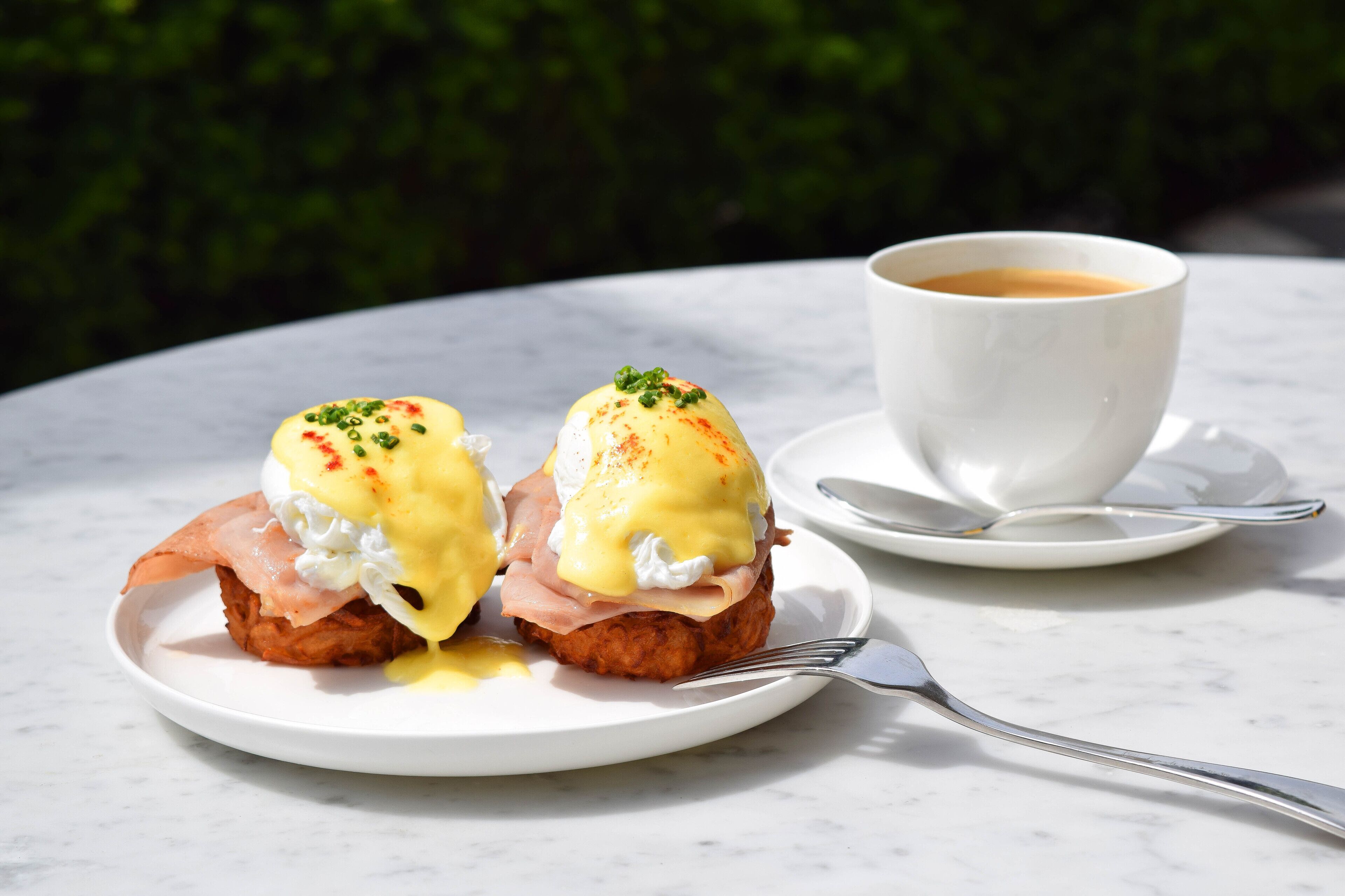 Two eggs benedict sandwiches on a white plate with a cup of coffee on a white saucer.