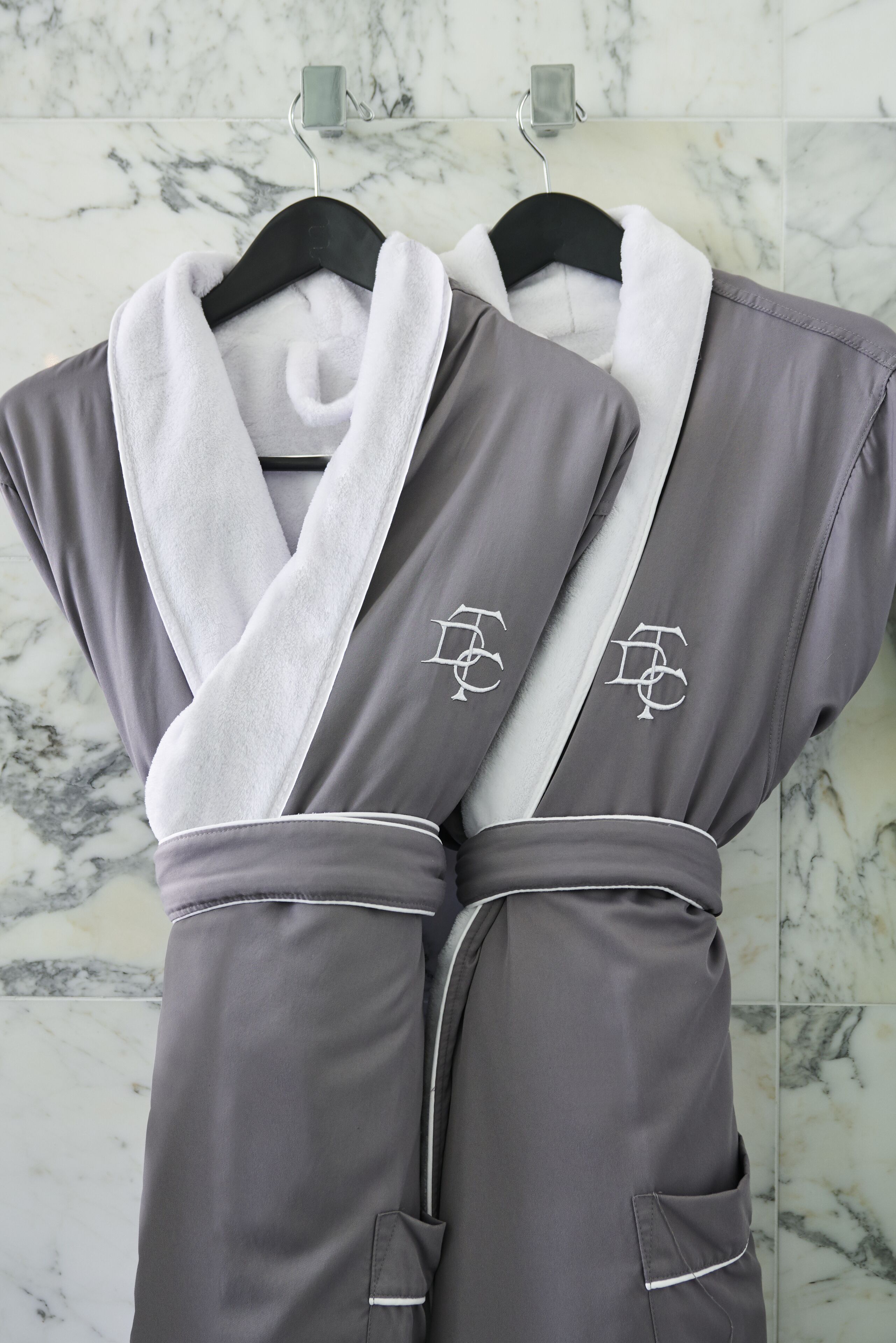 Two gray robes with white trim and embroidered logos on a rack against a marble wall.