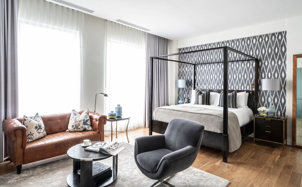 A modern hotel room with a leather sofa, a round coffee table, a grey armchair, a four-poster bed, and a patterned wallpaper.