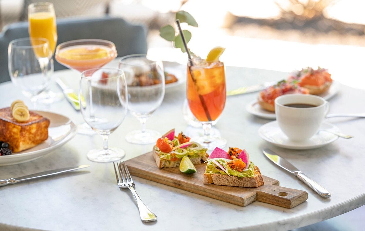 Ein Tisch reichhaltig mit Frühstücksangeboten gedeckt, darunter Toast, ein Smoothie und ein Cocktail, mit Blick ins Freie.