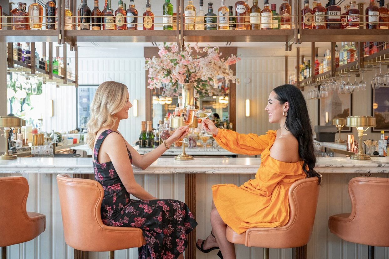 Two women seated at a bar with a variety of liquor bottles on shelves behind them.