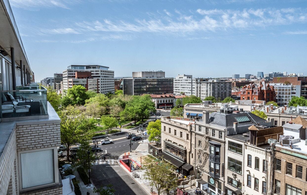 A cityscape with a mix of modern and historic buildings, a clear sky, and a street with cars and trees.