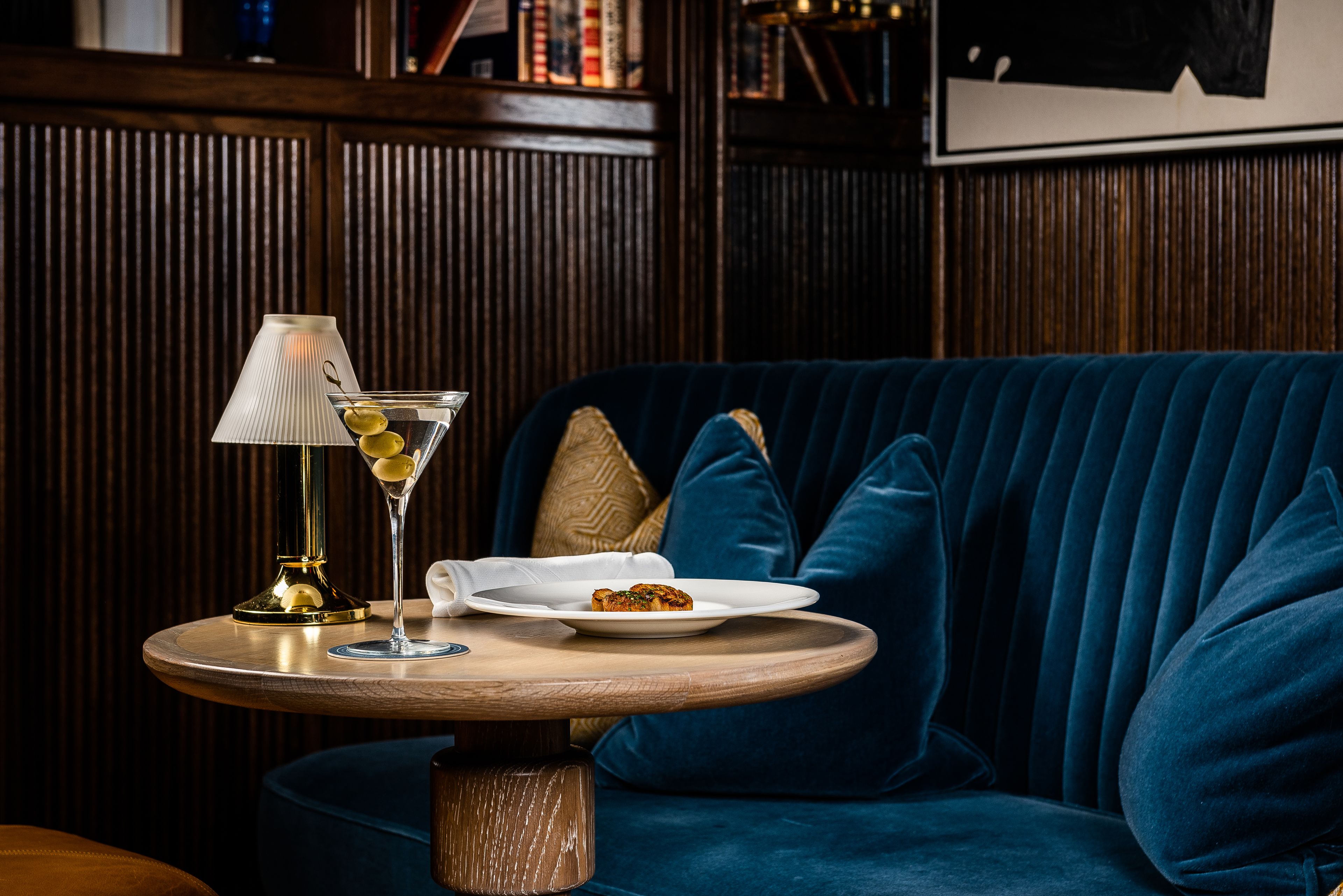 A round wooden table with a lamp, a martini glass, and a plate with a slice of cake.