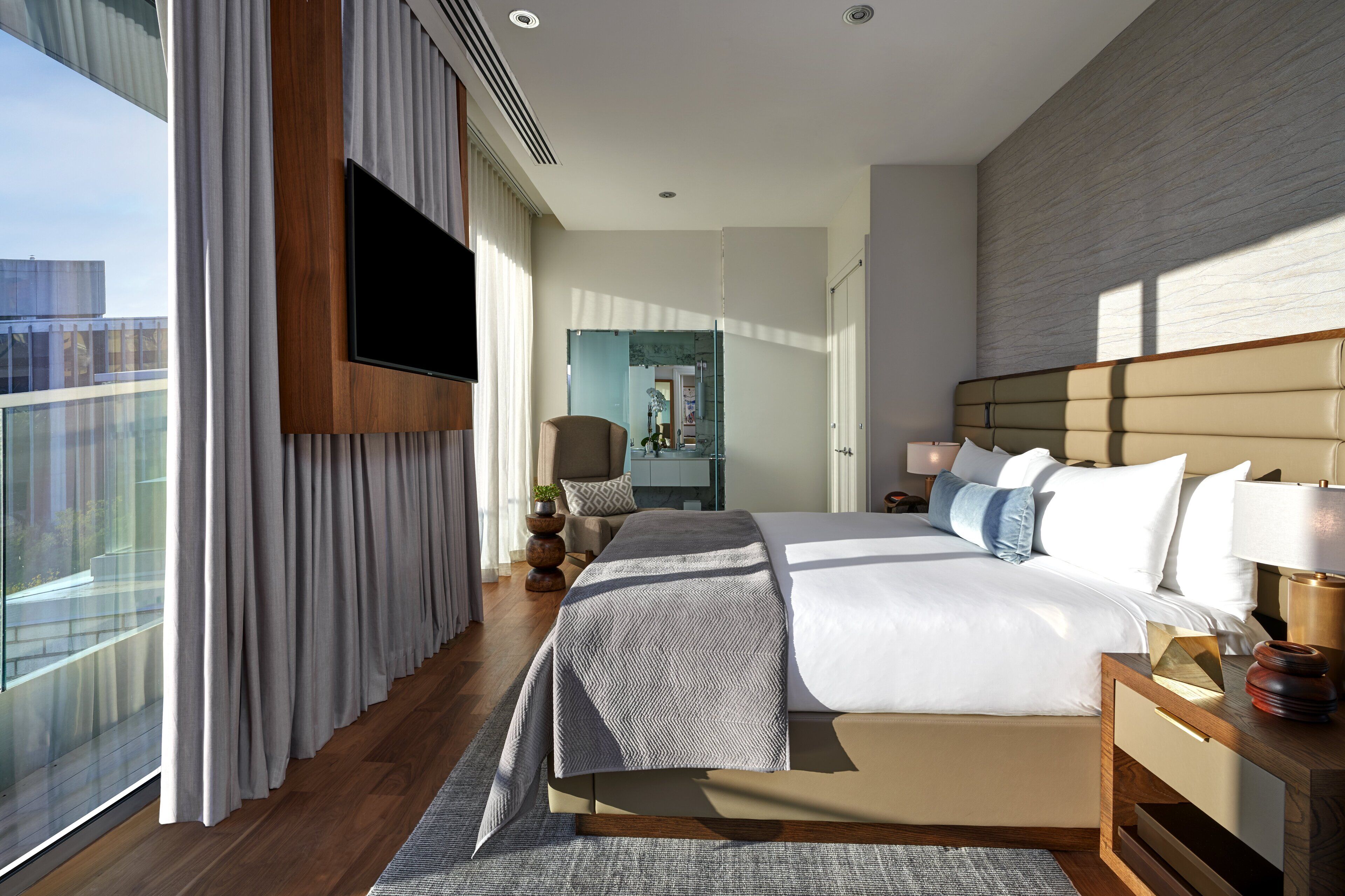 A modern hotel room with a large bed, a flat-screen TV mounted on the wall, a view of the city through large windows, and a wooden floor.