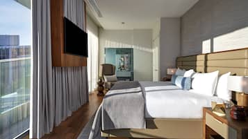 A modern hotel room with a large bed, a flat-screen TV mounted on the wall, a view of the city through large windows, and a wooden floor.