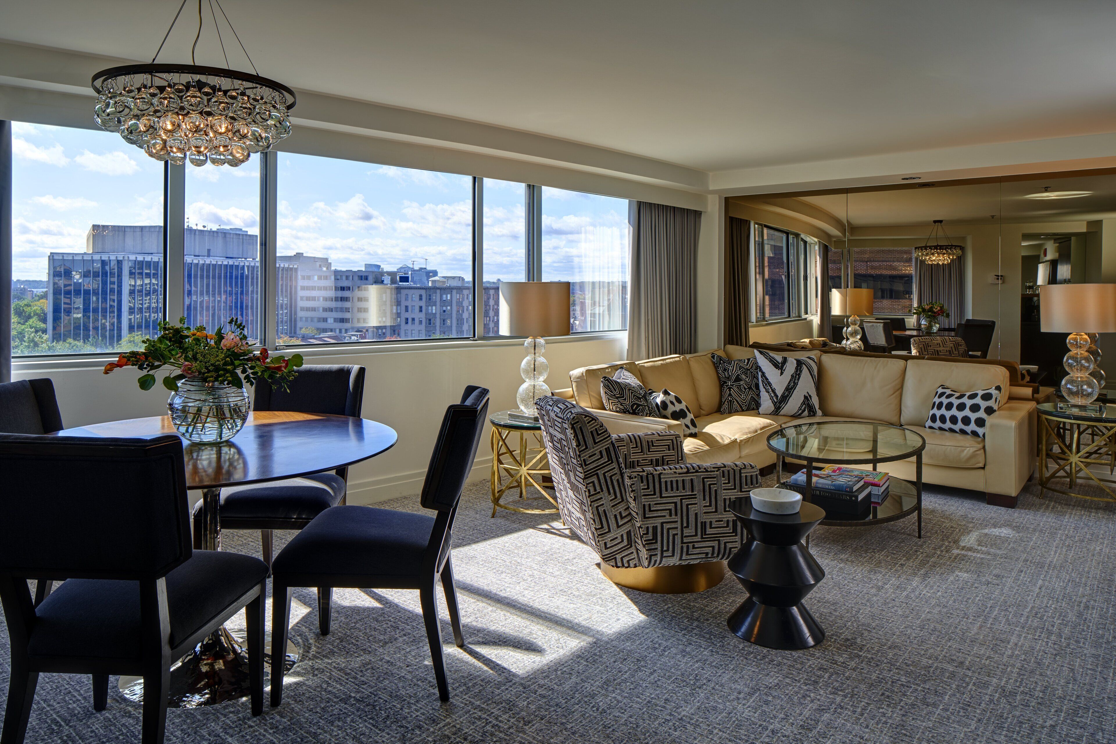 A modern hotel room with a large window, a dining area, a sofa set, and a small coffee table.