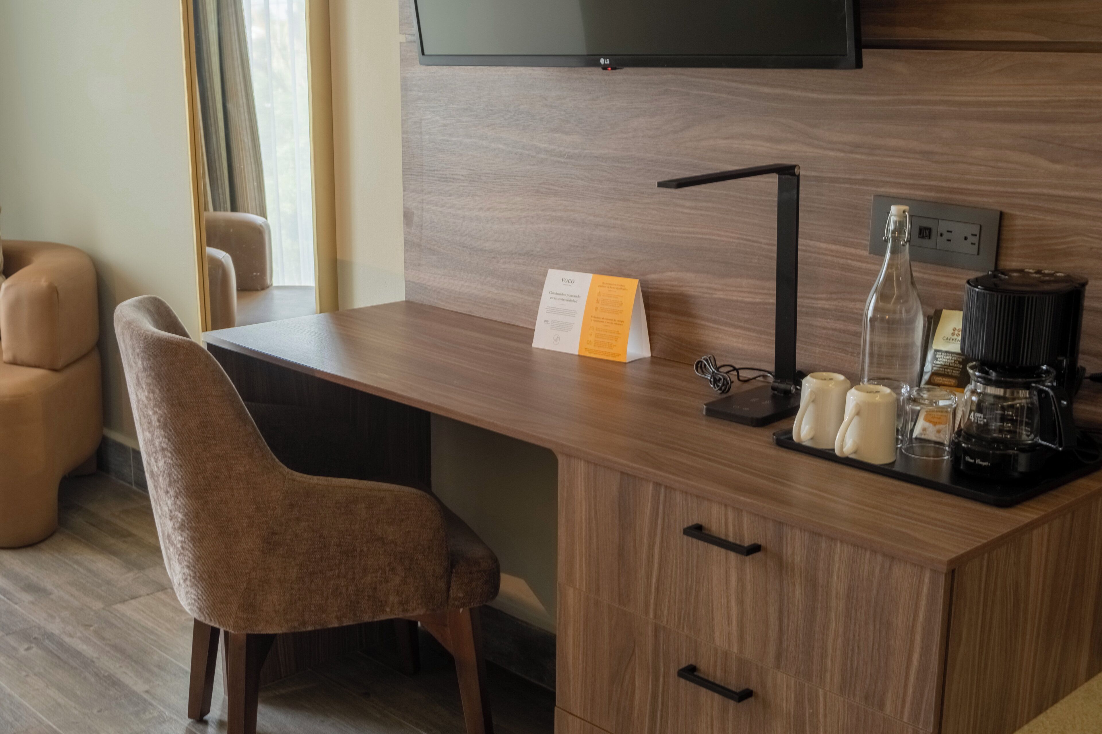 Een moderne hotelkamer met een houten bureau, een stoel, een aan de muur bevestigde televisie, een koffiezetapparaat en een fles.