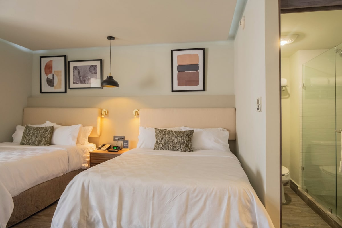 A hotel room with two beds, a nightstand with a phone, a framed abstract painting, a wall-mounted light, and a glass-enclosed shower.