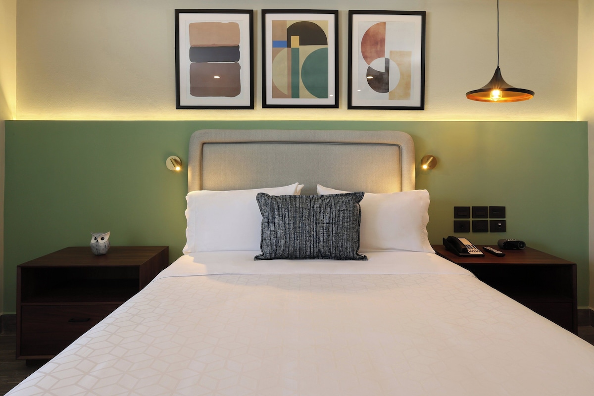 A neatly made bed with a patterned pillow, a bedside table with a phone and a lamp, and framed artwork on the wall.