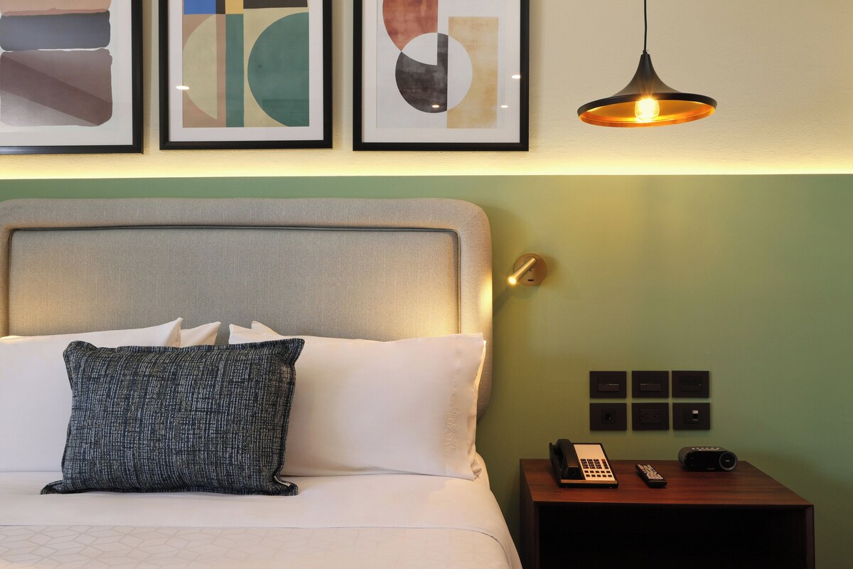 Een net opgemaakt bed met wit en grijs beddengoed, een grijs hoofdbord en een gestructureerd kussen. Een klein houten nachtkastje met een lamp, telefoon en afstandsbediening staat ernaast.