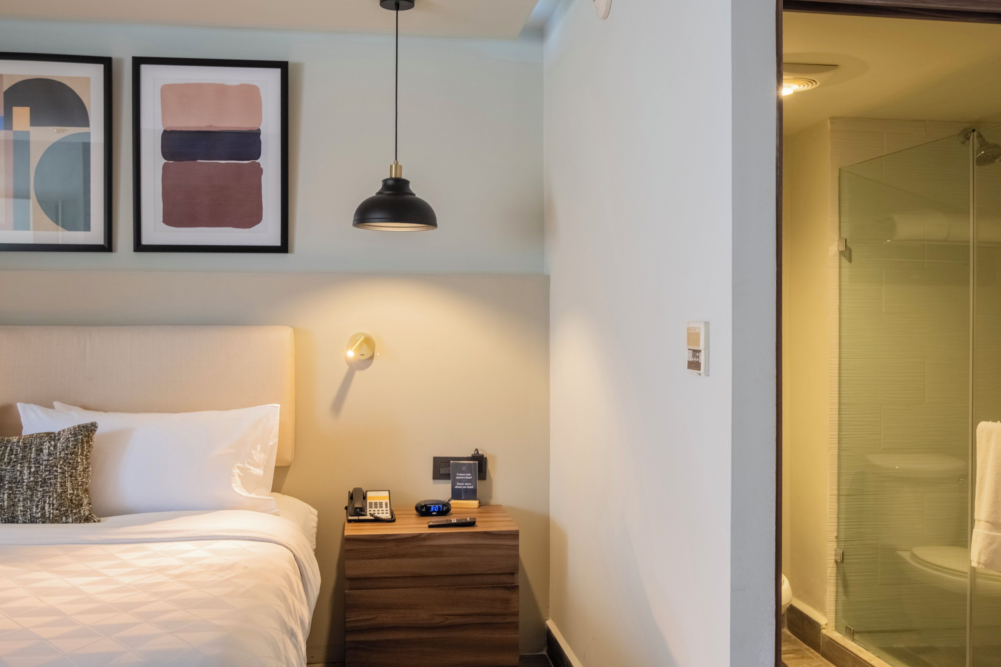 A hotel room with a bed, bedside table, lamp, and a glass-enclosed shower.