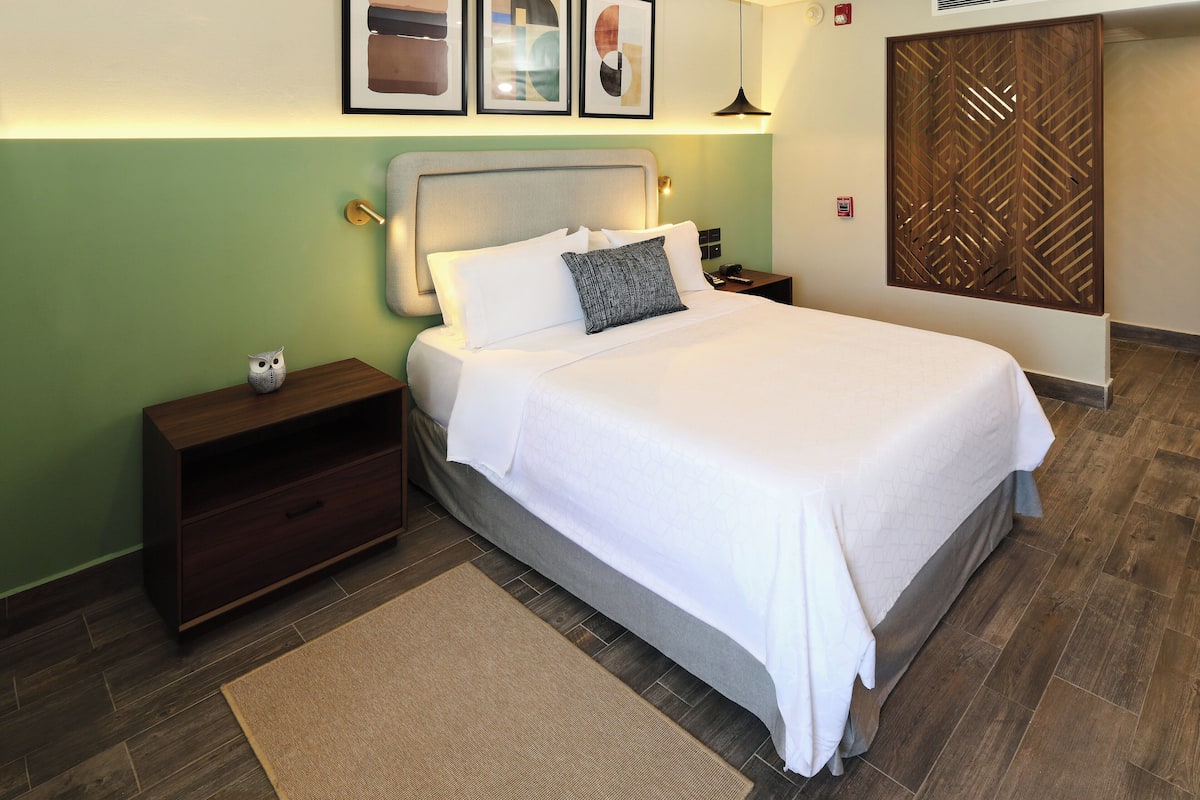 A neatly made bed with white linens, a wooden nightstand with a drawer, a small owl decoration, and a brown rug on a wooden floor.
