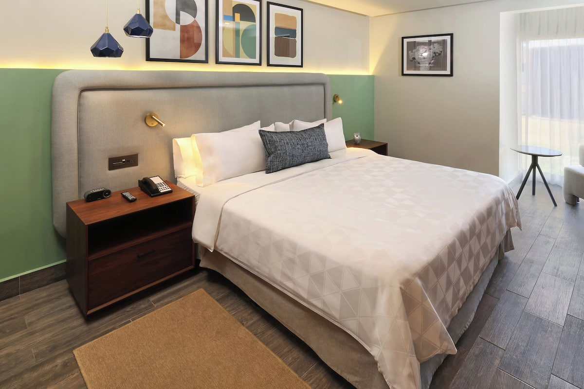A hotel room with a large bed, a bedside table with a phone and remote, a nightstand, a small table with a chair, and artwork on the walls.