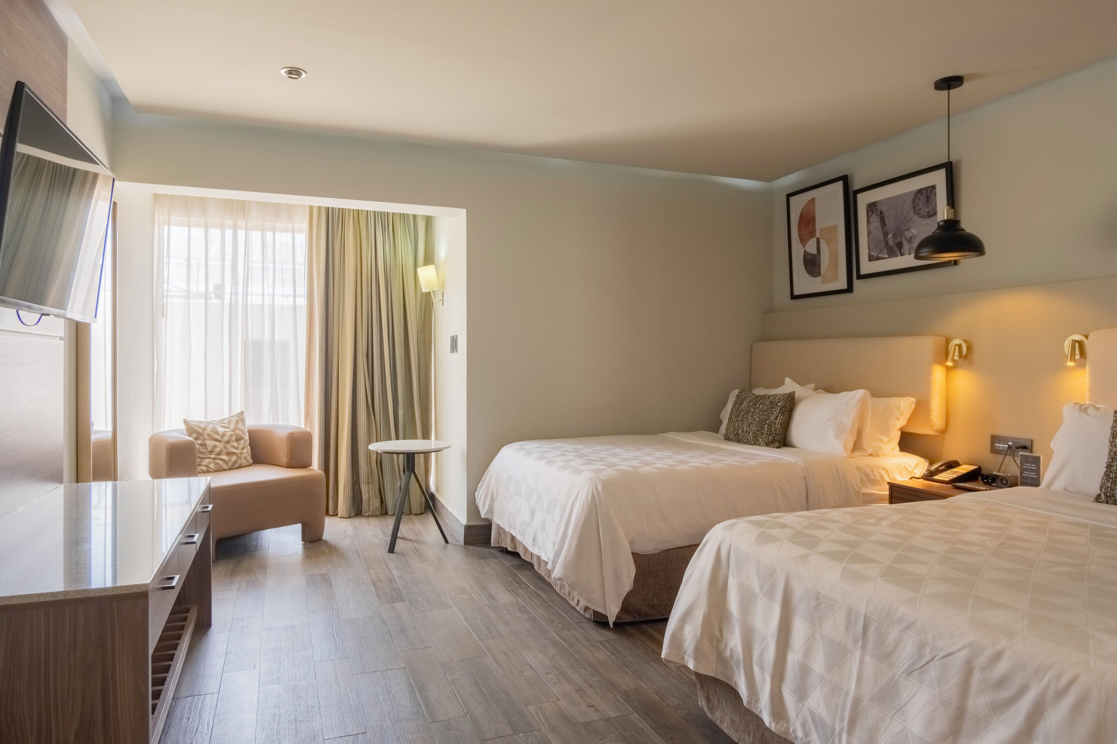 A hotel room with two beds, a TV, a desk, a chair, a small table, and a window with curtains.