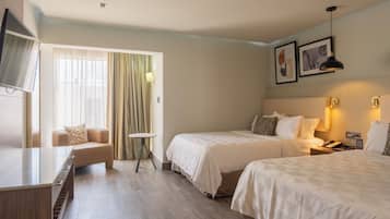 A hotel room with two beds, a TV, a desk, a chair, a small table, and a window with curtains.
