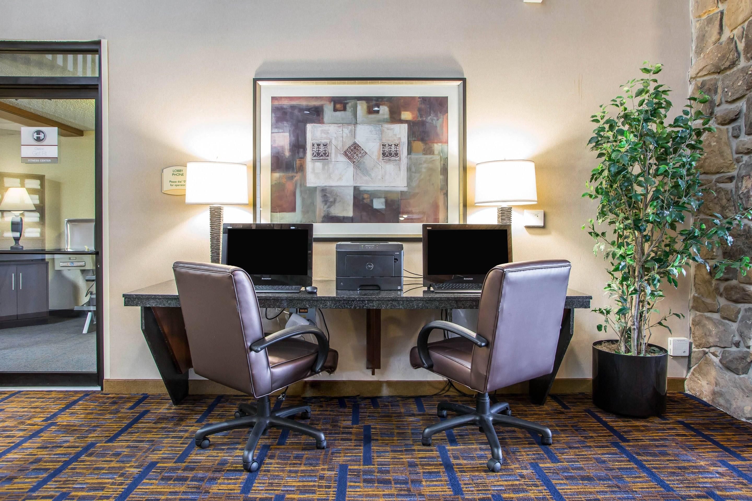 A hotel lobby with a reception desk, two computer workstations, two chairs, a potted plant, and a stone wall.
