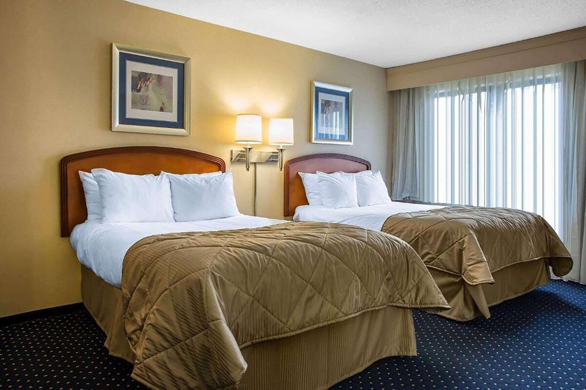 A hotel room with two beds, a large window with curtains, and wall-mounted lamps.