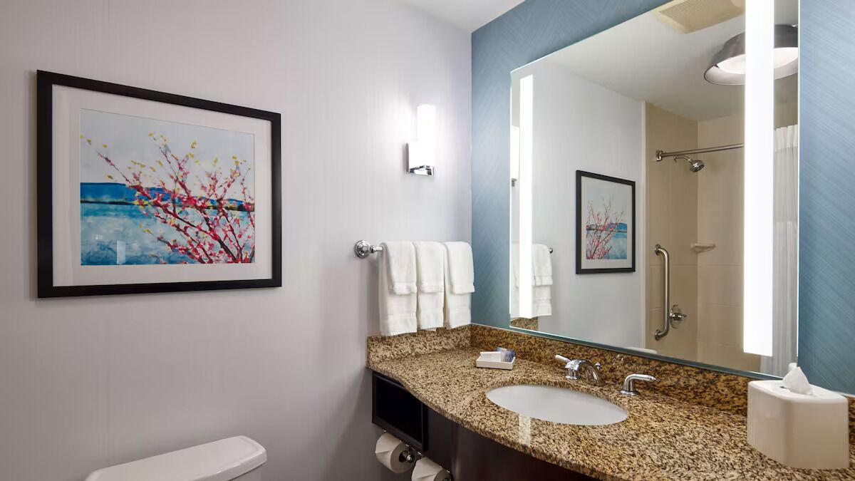 A modern bathroom with a granite countertop sink, a large mirror, and a framed painting on the wall.