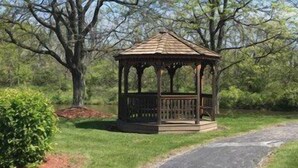 A gazebo with a wooden roof and open sides, surrounded by a paved path and lush greenery. - Clarion Inn (Merrillville)
