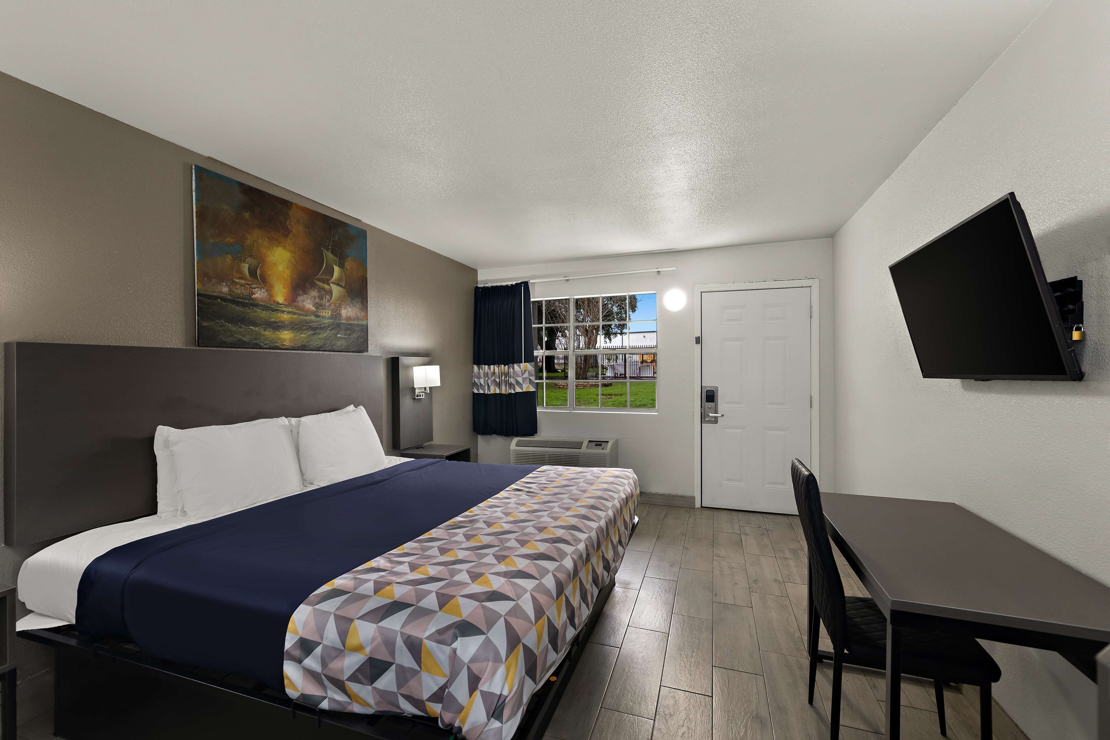 A hotel room with a bed, a desk, a chair, a TV mounted on the wall, and a painting above the bed.