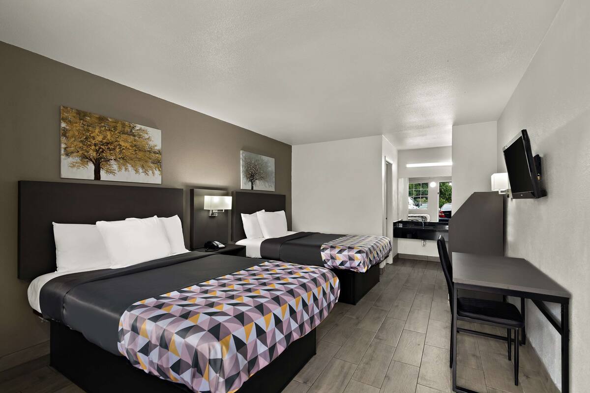 Habitación de hotel con dos camas, un escritorio y una silla. Hay televisores montados en la pared y una ventana con vistas al exterior.