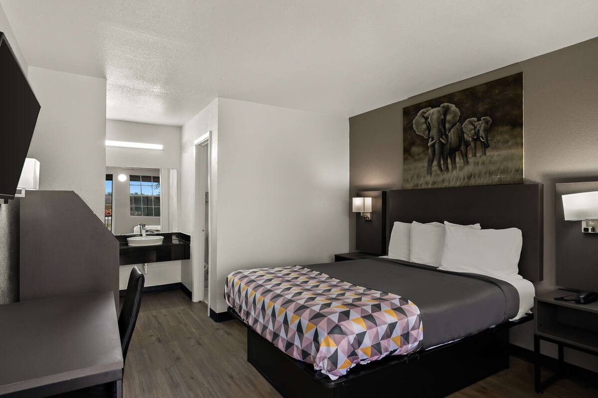 Una habitación de hotel moderna con una cama grande, un escritorio, una silla, un baño y un cuadro de elefantes en la pared.