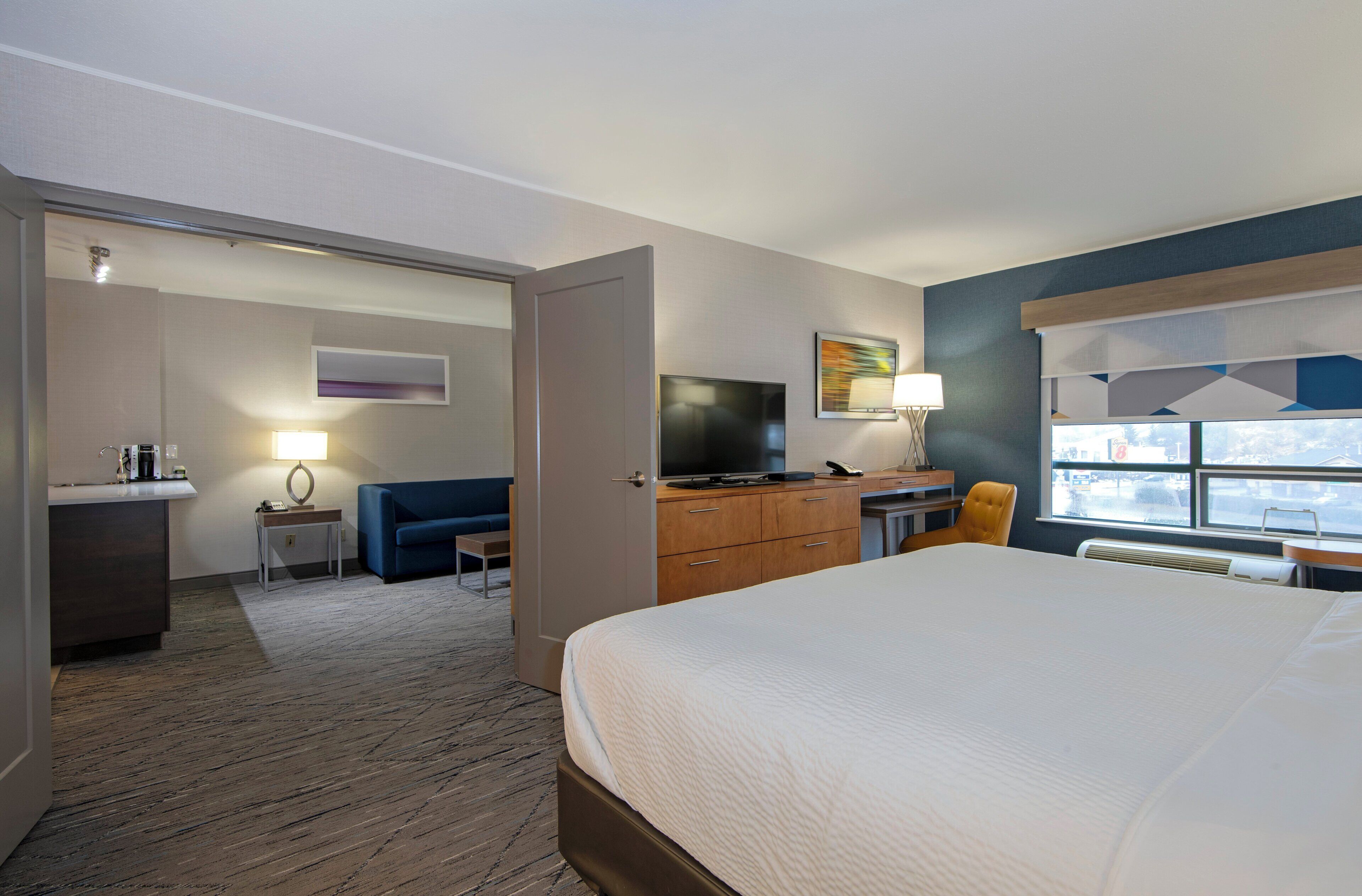 A hotel room with a bed, a desk with a TV, a sofa, a small table with a lamp, and a window with a view.