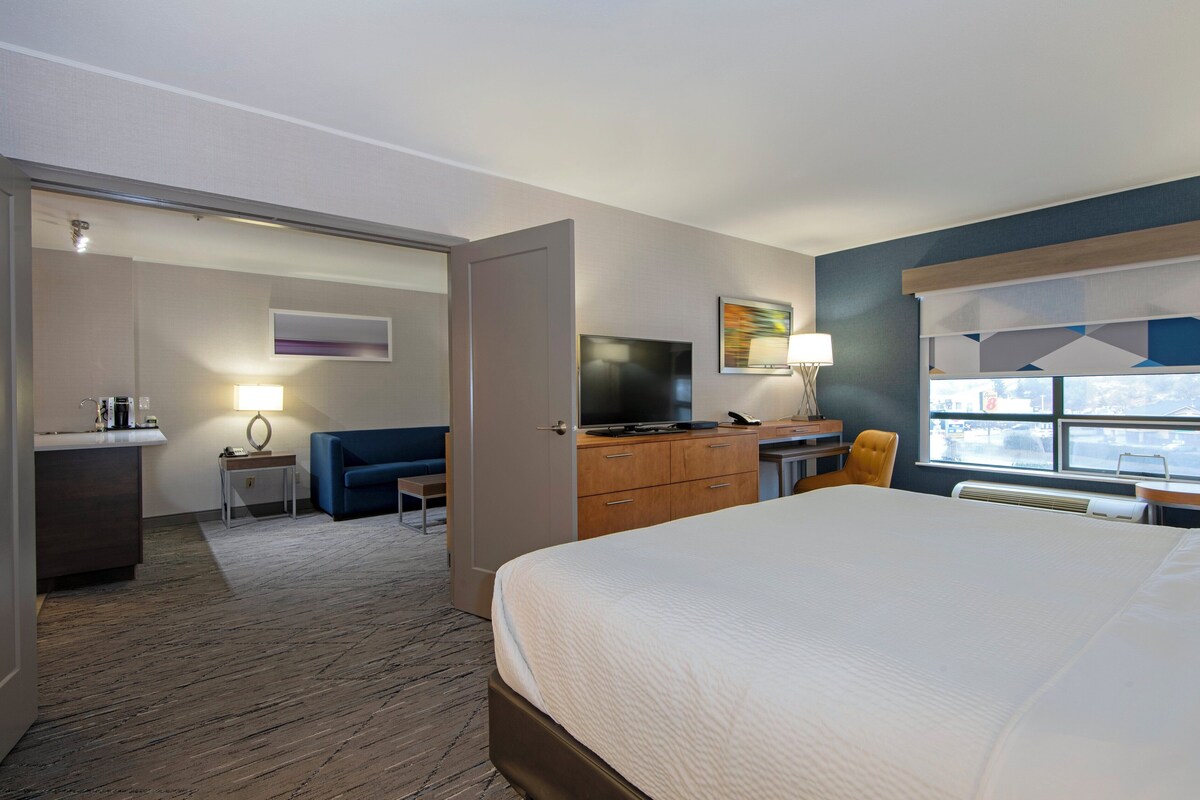 A hotel room with a bed, a desk with a TV, a sofa, a small table with a lamp, and a window with a view.