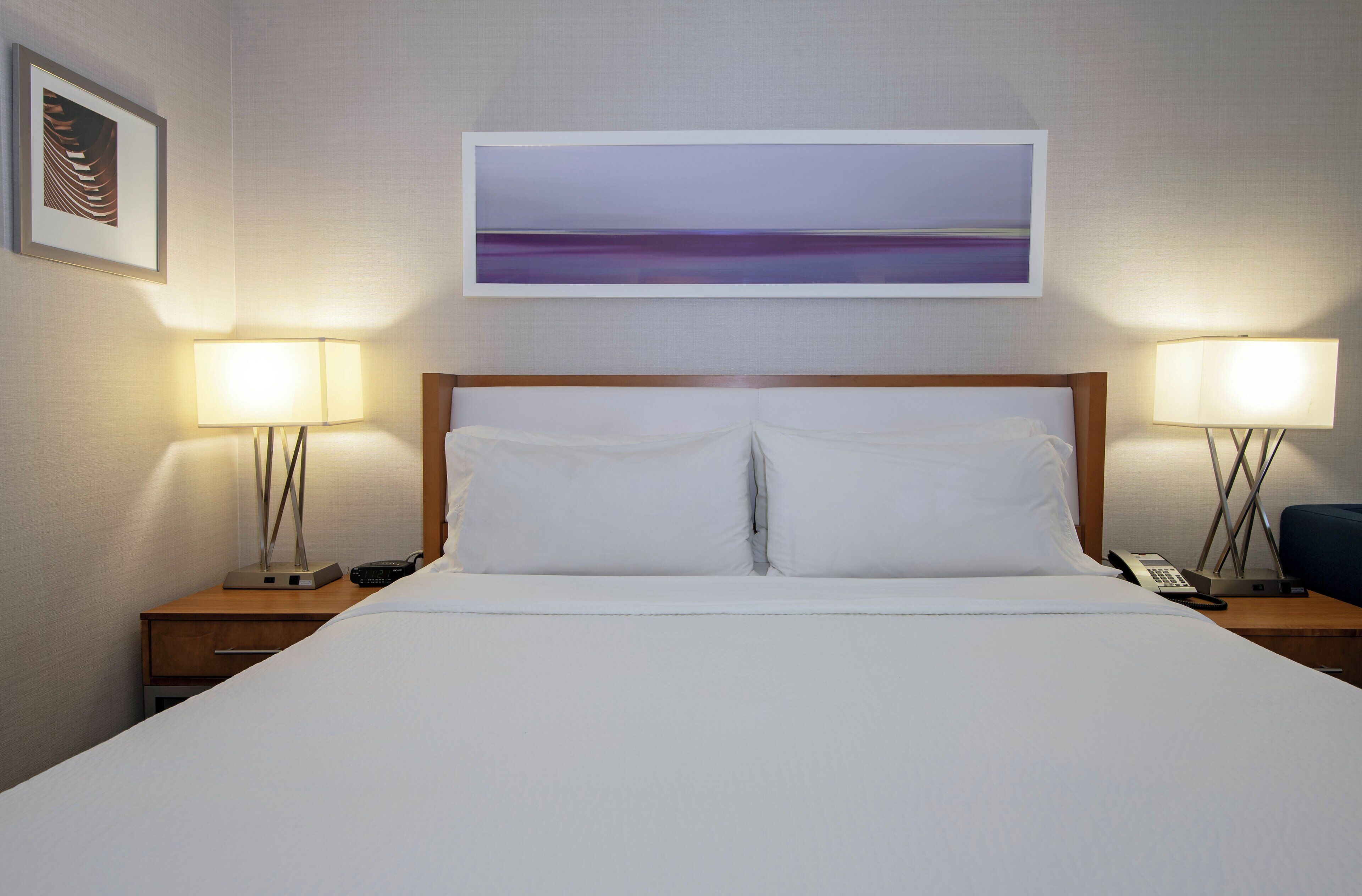 A hotel room with a large bed, two bedside tables with lamps, a painting above the bed, and a framed picture on the wall.