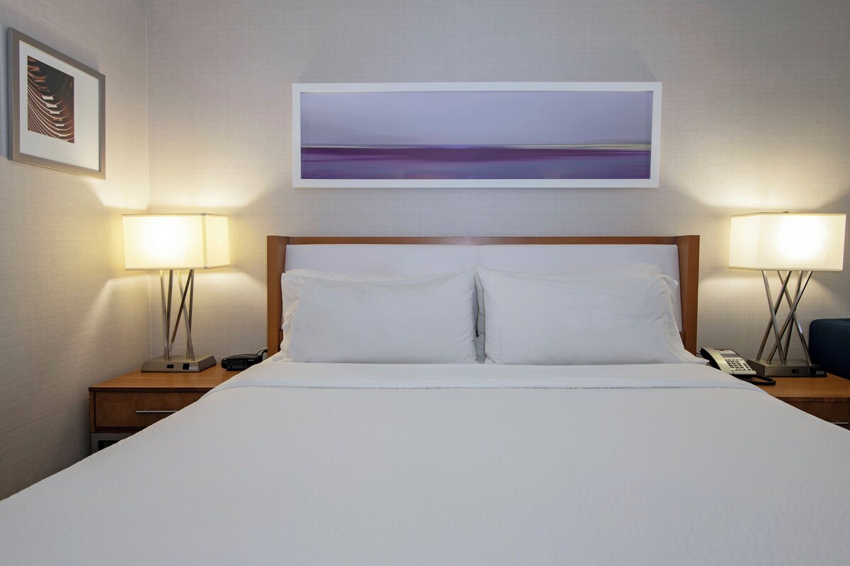 A hotel room with a large bed, two bedside tables with lamps, a painting above the bed, and a framed picture on the wall.