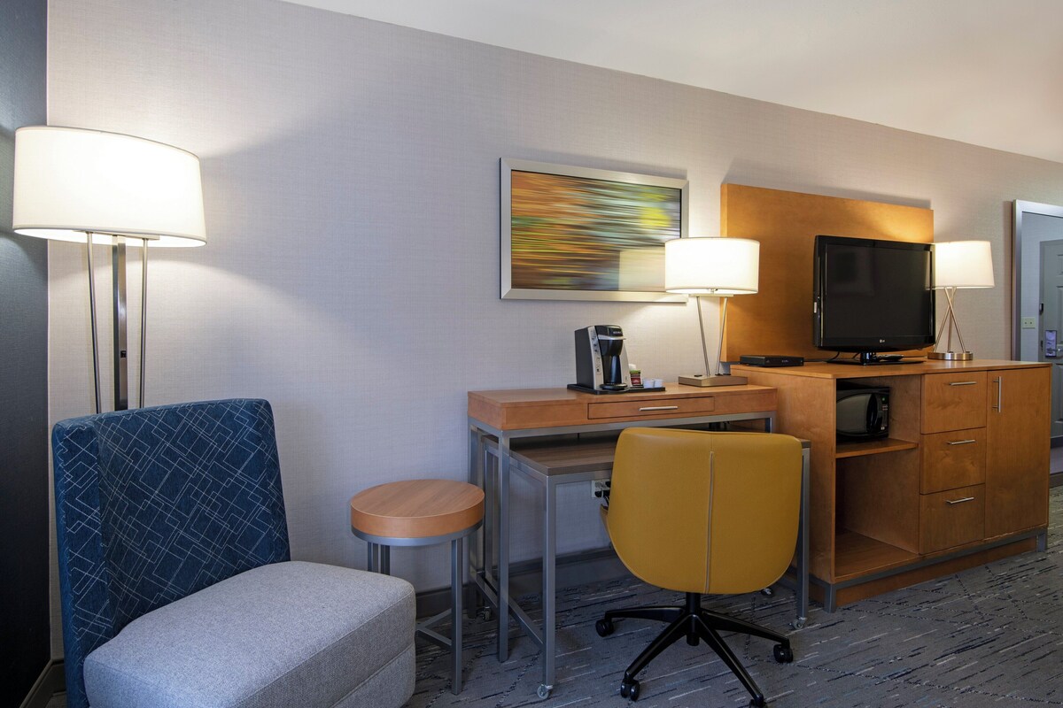 A hotel room with a desk, two chairs, a television, a coffee maker, and a painting on the wall.