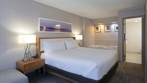 Habitación de hotel con una cama grande, lámparas de noche, una mesita de noche, un pequeño rincón de descanso y un baño visible a través de una puerta abierta.