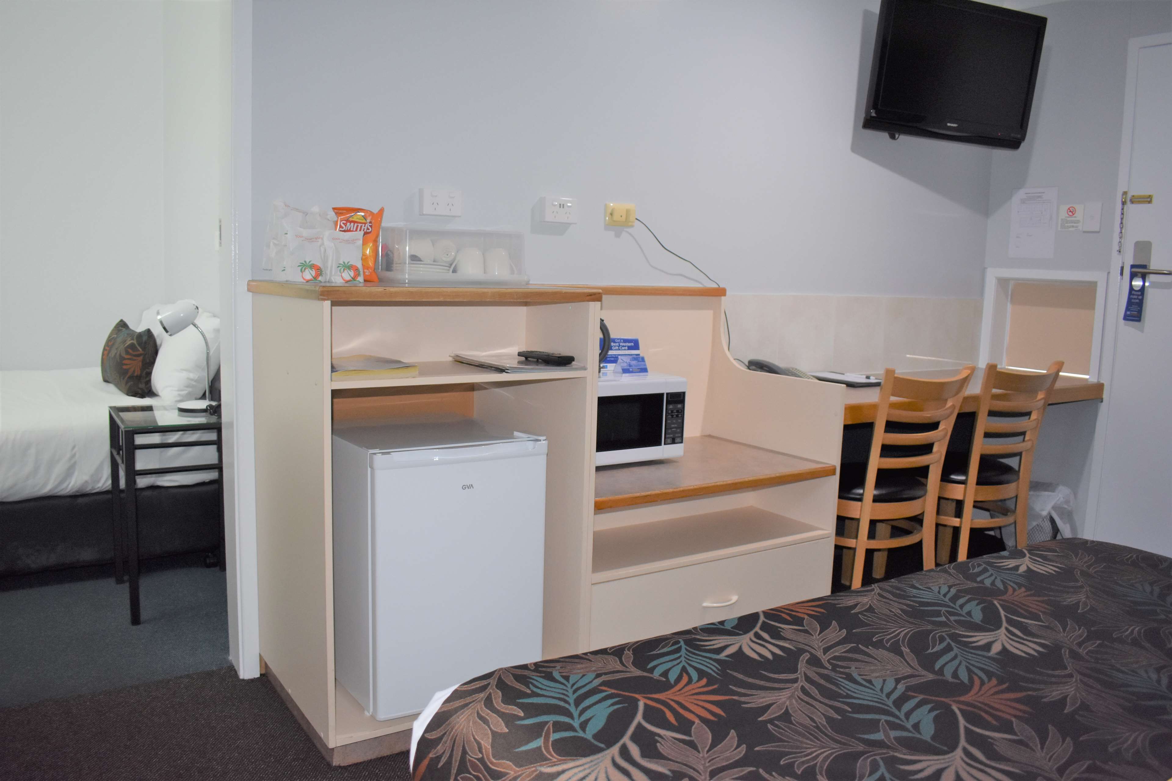 A compact hotel room with a bed, a small kitchenette, a microwave, a refrigerator, a desk, and a TV mounted on the wall.