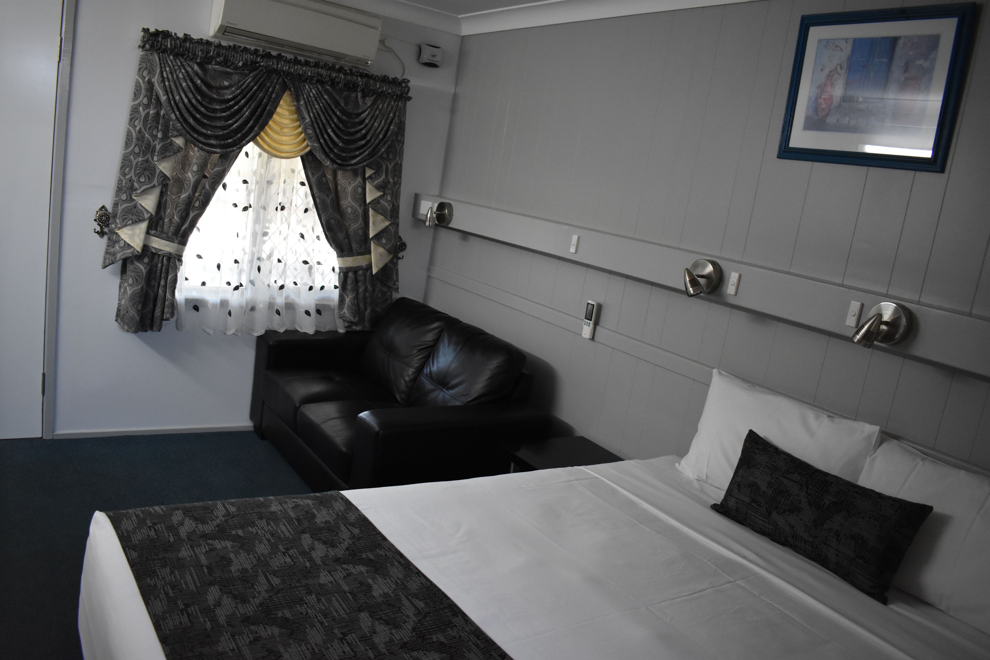 Ein Hotelzimmer mit einem Bett, einem schwarzen Ledersessel, einem Fenster mit gemusterten Vorhängen und einem gerahmten Bild an der Wand.