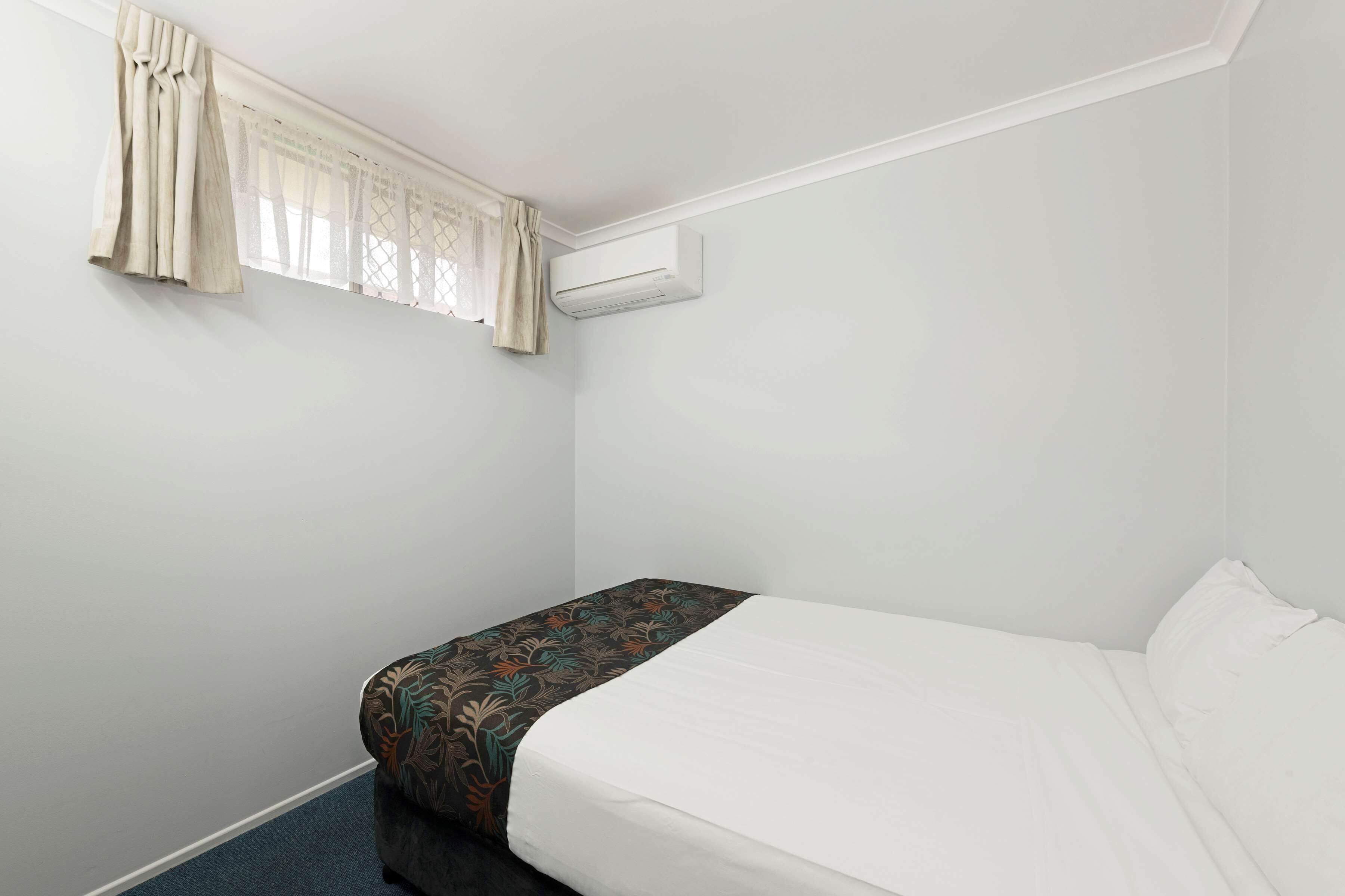 A small, single-bed room with a plain white wall, a window with sheer curtains, and an air conditioning unit mounted above the bed.