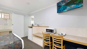 A hotel room with a bed, a desk with a microwave, two chairs, a TV mounted on the wall, and a small trash bin.