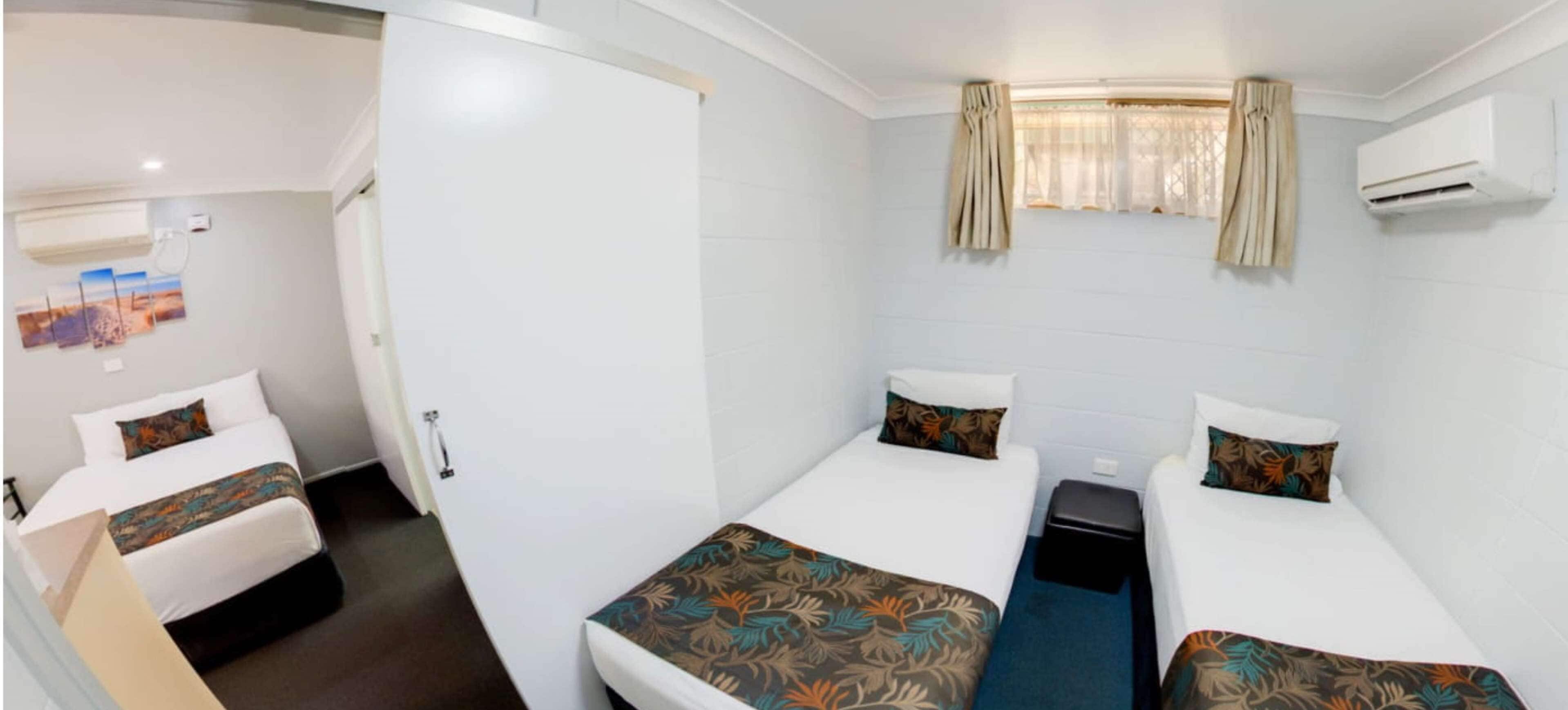 A hotel room with two single beds, a small table, a wall-mounted air conditioner, and a window with curtains.